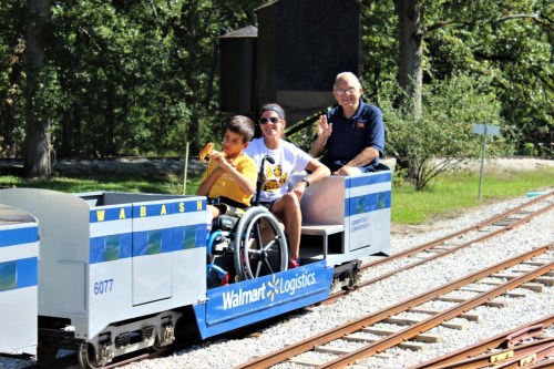 A boy in a wheelchair and two adults enjoy a ride on a small train, waving and smiling in a park setting.