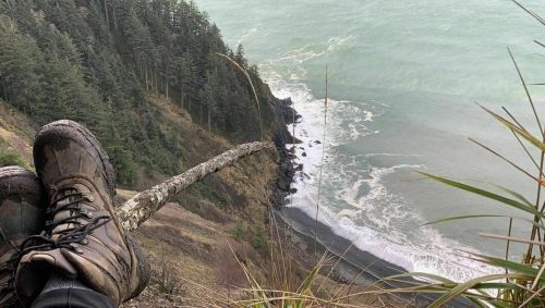 A hiker's muddy boots resting on a log overlooking a rocky coastline and waves crashing below.