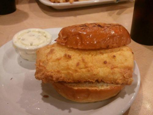 A fried fish sandwich on a toasted bun, served with a small cup of tartar sauce on a white plate.