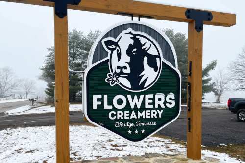 Sign for Flowers Creamery in Ethridge, Tennessee, featuring a cow graphic and snowy surroundings.