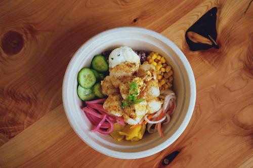 A colorful bowl of rice topped with fried fish, cucumbers, corn, pickled onions, and various vegetables.