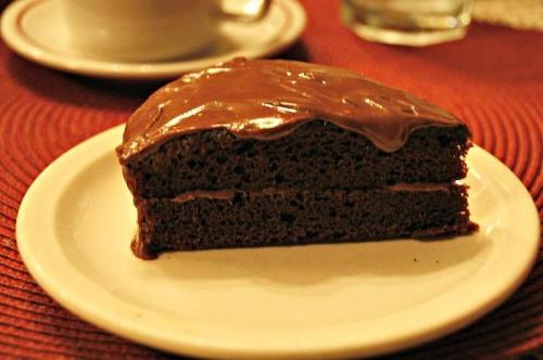 A slice of chocolate cake with smooth frosting on a white plate, set on a textured red tablecloth.