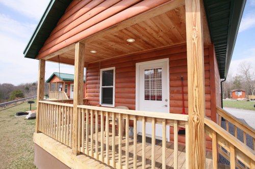 A wooden porch with a swing, leading to a log cabin with a white door and windows, surrounded by greenery.