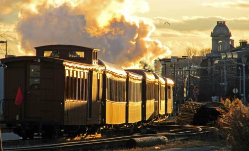 A vintage train glows in the sunset, with steam rising and a historic building in the background.