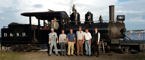 A group of seven people poses in front of a vintage steam locomotive labeled "B. & S. R." by a waterfront.
