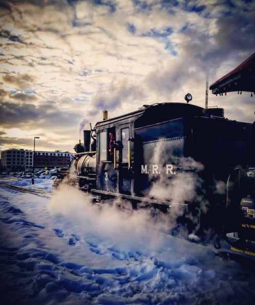 A vintage steam train billows smoke as it sits on snowy tracks under a cloudy sky.