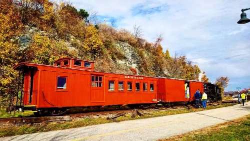 A bright red train car on a scenic railway track, surrounded by autumn foliage and a cloudy sky.