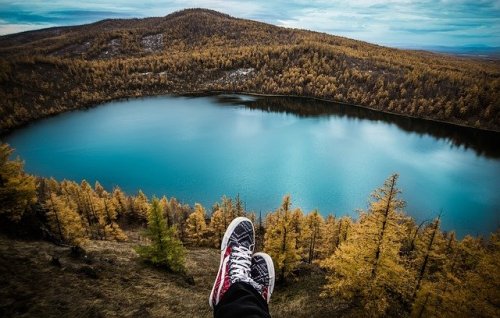 A person’s feet in sneakers resting on a ledge overlooking a serene blue lake surrounded by autumn trees.