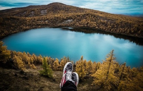 A person’s feet in sneakers resting on a ledge overlooking a serene blue lake surrounded by autumn-colored trees.