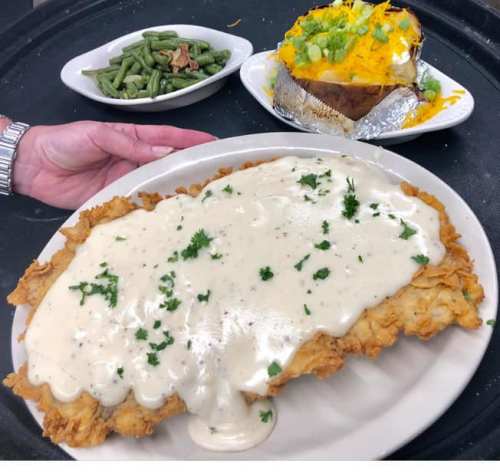 A large plate of chicken fried steak smothered in gravy, served with a baked potato and green beans on the side.