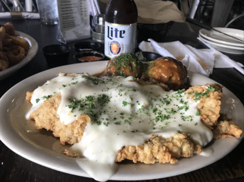 A plate of fried chicken topped with white gravy and parsley, accompanied by meatballs and a bottle of beer.