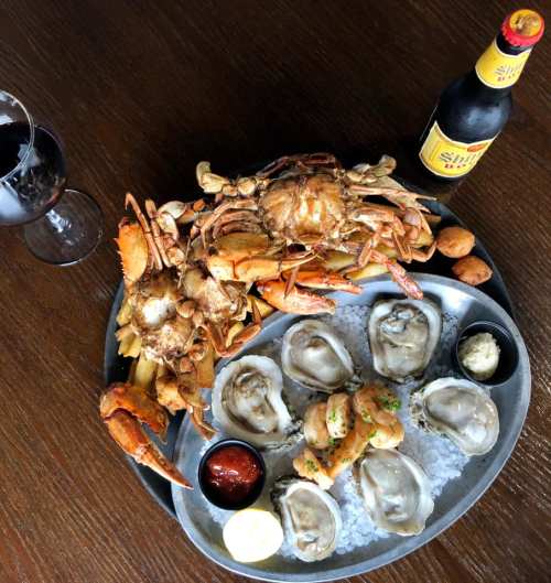 A seafood platter with crabs, oysters, shrimp, sauces, and a beer, served on a metal tray with a glass of wine.