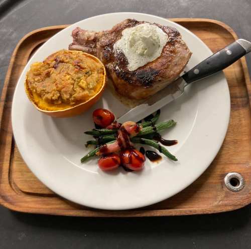 A plated meal featuring a grilled pork chop with herb butter, stuffed squash, roasted tomatoes, and asparagus on a wooden tray.
