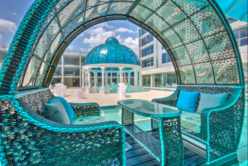 A stylish outdoor seating area with turquoise wicker furniture, overlooking a dome structure and a sunny pool area.