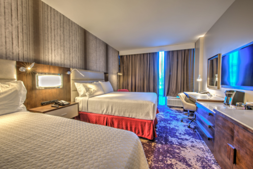 A modern hotel room featuring two beds, a desk, and large windows with curtains, illuminated by soft lighting.