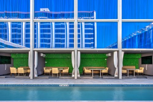 Modern poolside view featuring glass walls, green accents, and dining areas reflecting in the water.