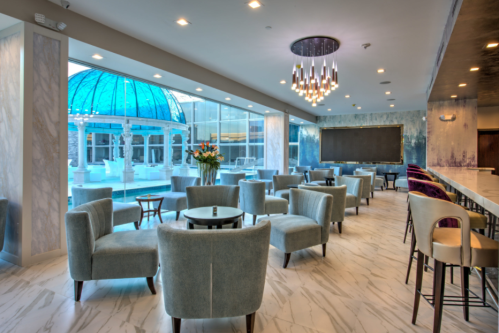 Modern lounge area with plush chairs, a bar, and a glass dome ceiling overlooking a pool. Elegant lighting and decor.