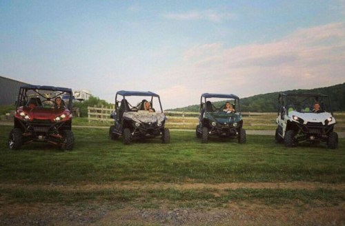 Four off-road vehicles parked on grass, with people sitting inside, against a backdrop of trees and a cloudy sky.