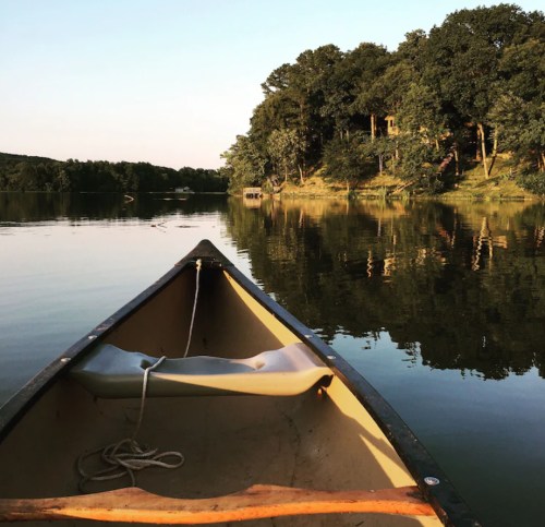 A canoe on calm water, reflecting trees and a clear sky, with a distant shoreline visible.