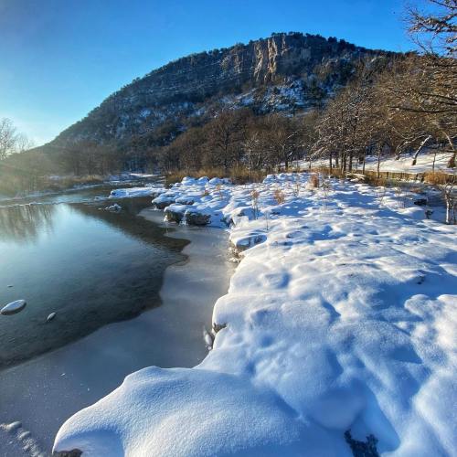 A serene winter landscape featuring a snowy shore, calm water, and a mountain in the background under a clear blue sky.