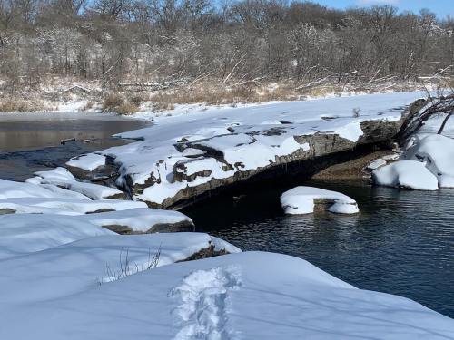 A snowy landscape with a rocky outcrop over a calm river, surrounded by trees and a clear blue sky.