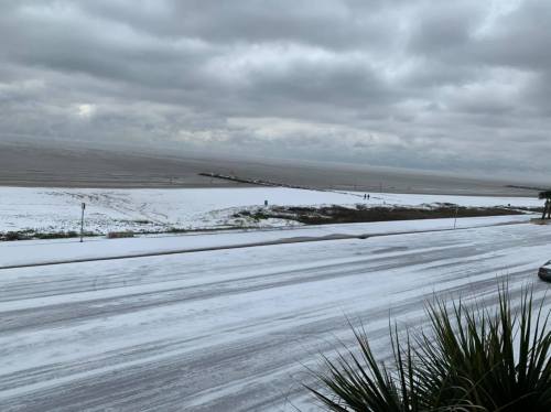 A snowy beach scene with a cloudy sky, featuring a road lined with snow and a pier in the distance.