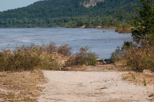 A sandy path leads to a riverbank, surrounded by greenery and hills in the background under a clear sky.