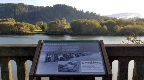 View of a river with lush greenery, featuring an informational sign about the area's history and landscape.