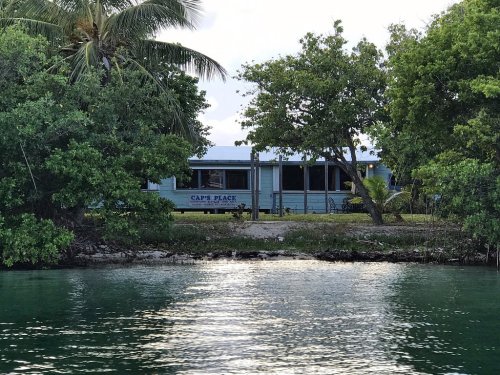 A blue building named "Cap's Place" surrounded by greenery, viewed from a calm waterway.