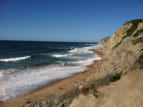 A scenic view of a sandy beach with gentle waves, rocky cliffs, and a clear blue sky.