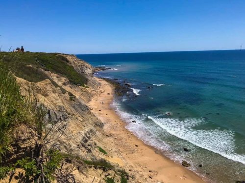 A scenic view of a sandy beach with rocky shores, cliffs, and a clear blue sky over the ocean.