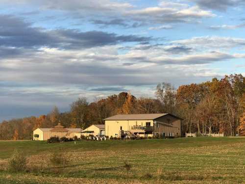 A large beige building surrounded by trees with autumn foliage, set against a cloudy sky and open fields.