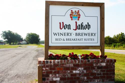 Sign for Von Jakob Winery, Brewery, and Bed & Breakfast Suites, featuring flowers and a rural landscape.