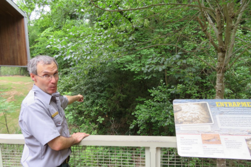 A park ranger gestures towards a wooded area while standing next to an informational sign about entrapment.