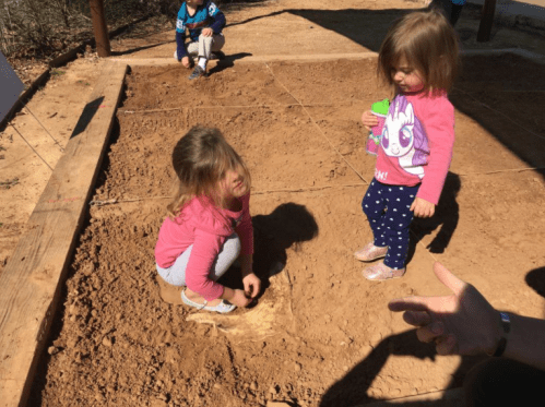 Two young girls play in a garden bed, one kneeling in the dirt while the other stands nearby holding a drink.
