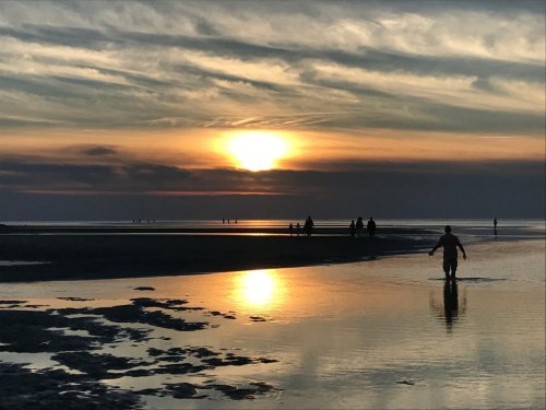 A sunset over a calm beach, with silhouettes of people walking along the shore and reflections in the water.