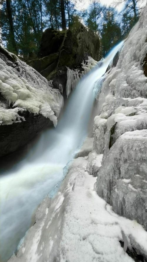 A frozen waterfall cascading over icy rocks, surrounded by trees in a winter landscape.