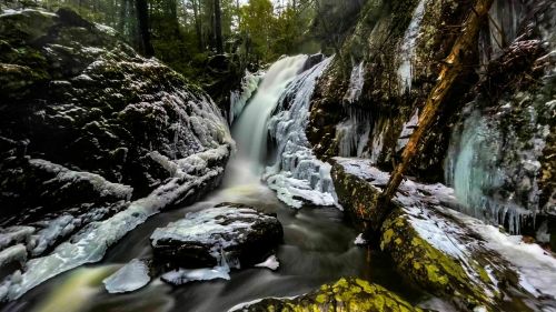 A serene waterfall cascades over icy rocks, surrounded by lush greenery and frozen formations in a tranquil forest setting.