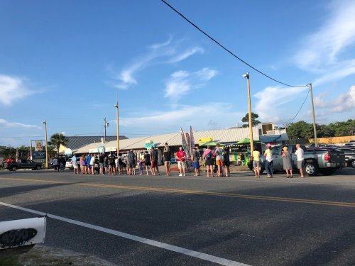 A long line of people waiting outside a restaurant on a sunny day, with cars parked along the road.