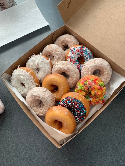 A box of assorted donuts, including glazed, frosted, and sprinkled varieties, with some topped with coconut and cereal.