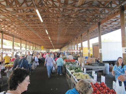A bustling farmers market with people browsing stalls filled with fresh produce under a wooden roof.