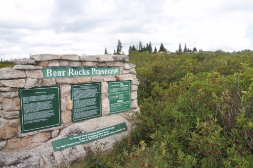 Sign at Bear Rocks Preserve with informational plaques, surrounded by green vegetation and trees under a cloudy sky.