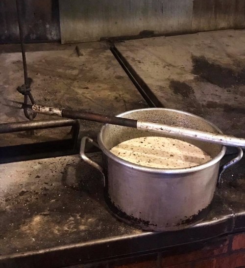 A metal pot with a creamy substance sits on a dirty, rustic countertop, alongside a long, metal stirring rod.
