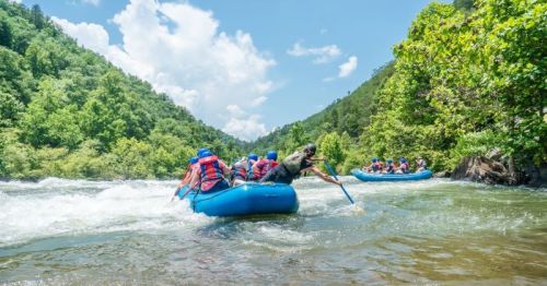 A group of people in blue rafts navigating a river surrounded by lush green hills and a bright blue sky.