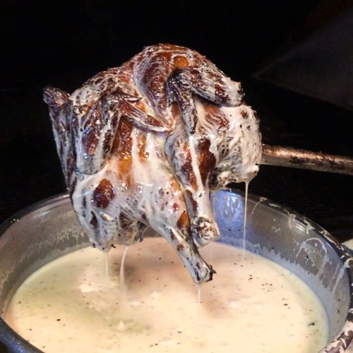 A roasted chicken on a spit, dripping with creamy sauce, being lifted from a pot.