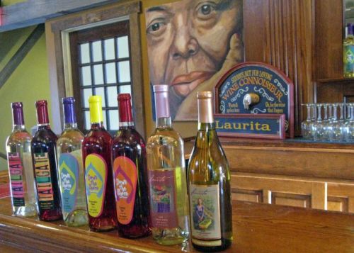 A wooden bar displays various colorful bottles of wine, with a large portrait of a person in the background.