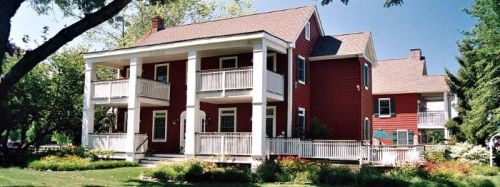 A red two-story house with white columns, balconies, and a landscaped yard under a clear blue sky.