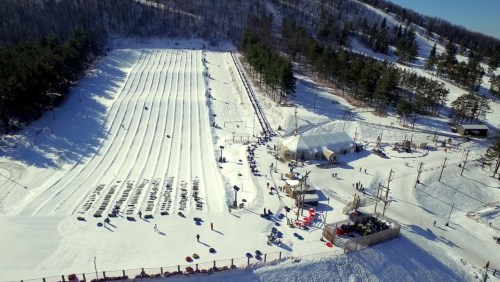 A snowy hill with multiple tubing lanes, a crowd of people, and a large tent in a winter landscape.