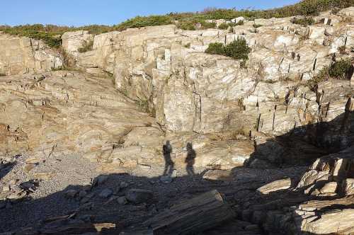 Two shadows cast on rocky terrain, with a clear blue sky above and greenery in the background.