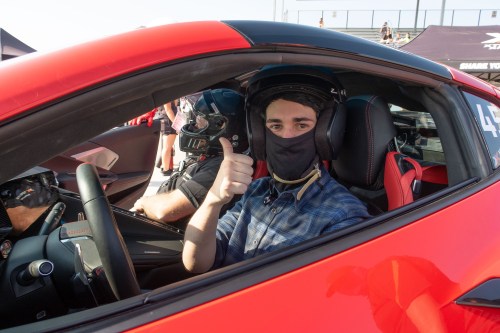 A driver in a red sports car gives a thumbs up, wearing a helmet and face mask, with another person in the background.